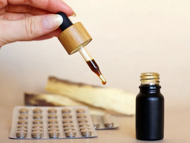 Hand hält eine Pipette mit braunem Tropfen über einer Flasche, daneben ein Blister mit Tabletten