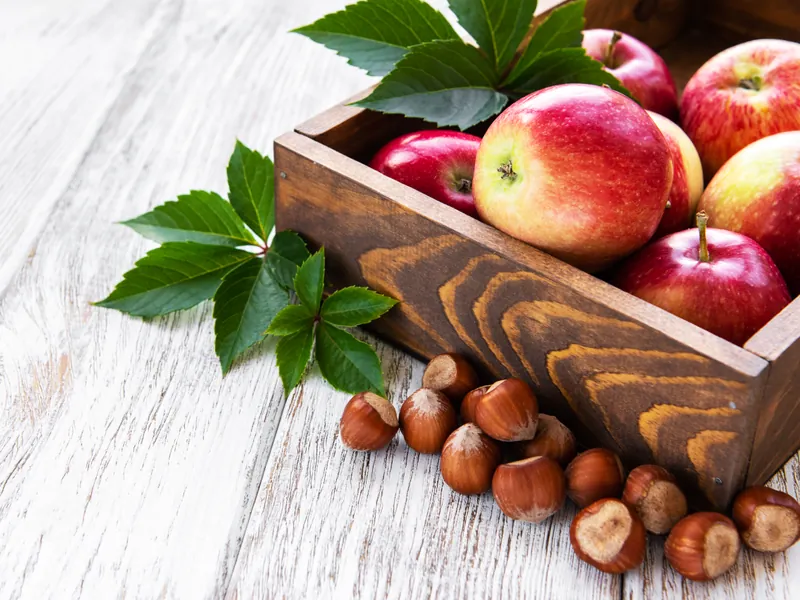 Rote Äpfel in einer Holzkiste, davor ganze Haselnüsse auf hellem Holztisch