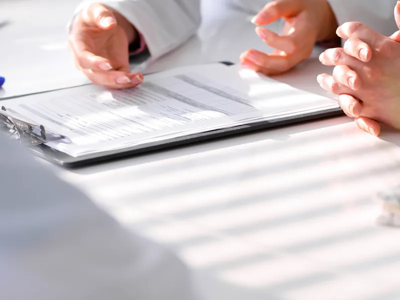 Hände von Ärztin und Patientin an einem Tisch mit Unterlagen im Tageslicht